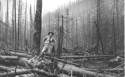 黑白相片显示一名男子身着领带站在森林中,周围环绕着烧焦日志